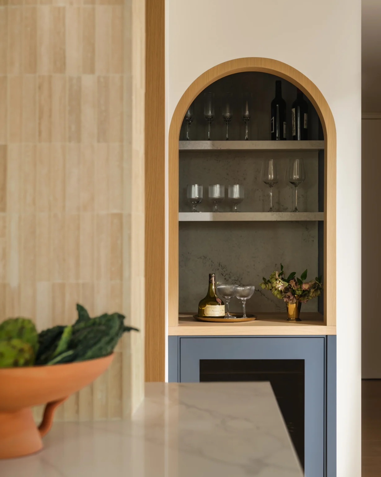 Cool arched bar nook interior design in a Manhattan apartment with warm wood trim, glassware shelves, wine bottles, and blue cabinetry