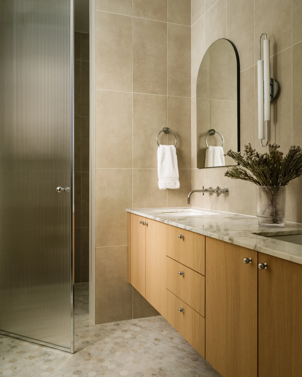 Modern primary bathroom interior design in a Manhattan apartment with oak vanity, marble countertop, arched mirror, neutral tile, and glass shower