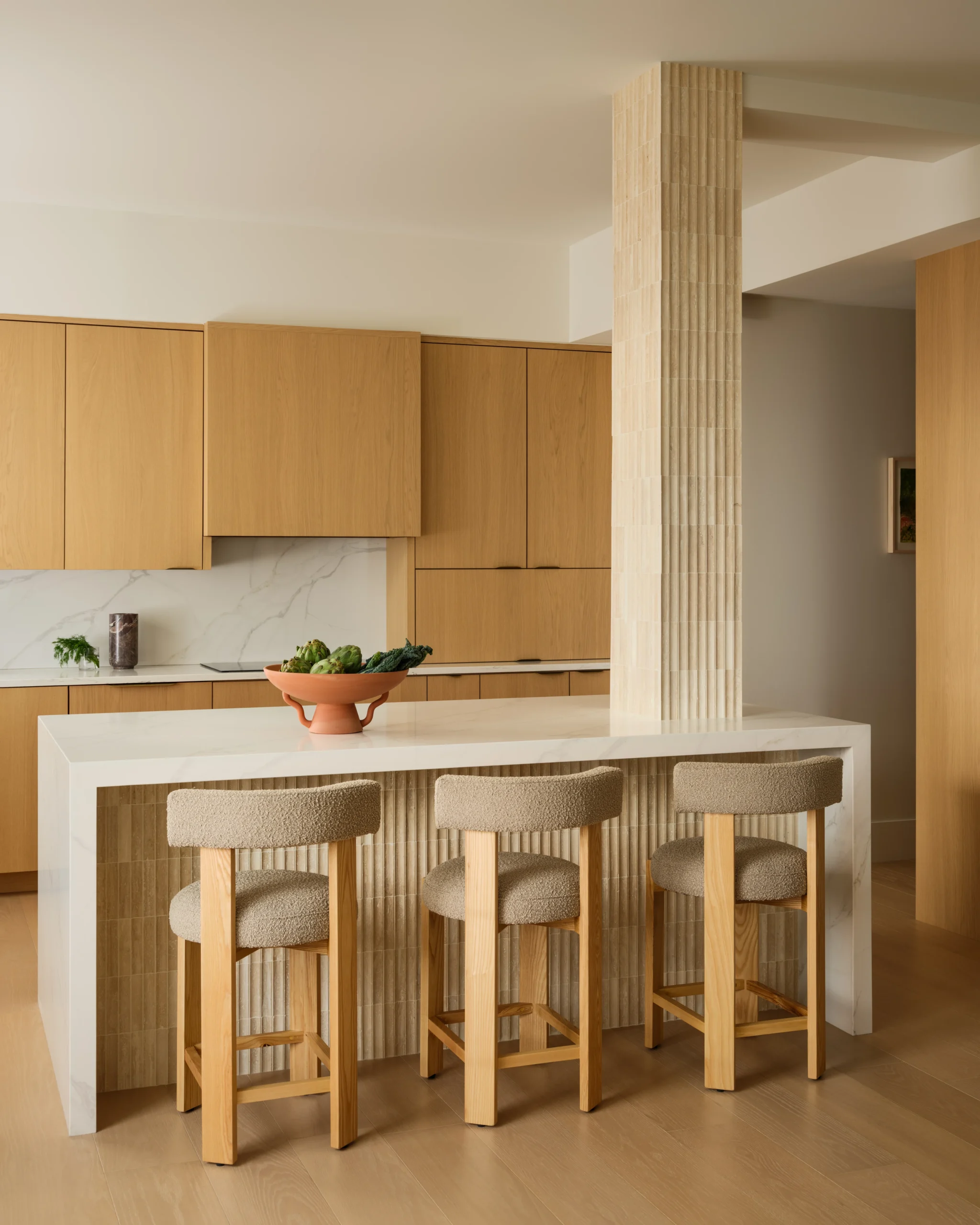 Modern organic kitchen interior design in a Manhattan apartment with oak cabinets, marble island, textured column, and upholstered wood stools