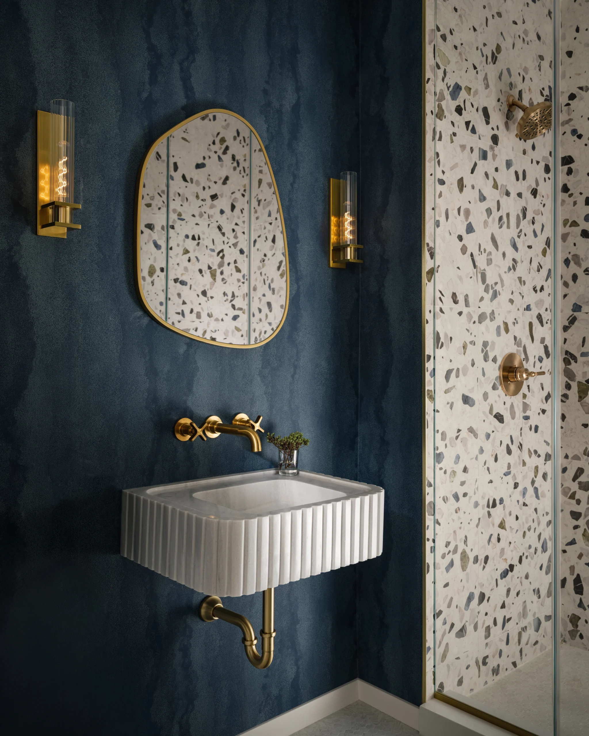 Moody powder room interior design in a NYC apartment with deep blue walls, brass lighting, fluted sink, terrazzo shower tile, and gold accents