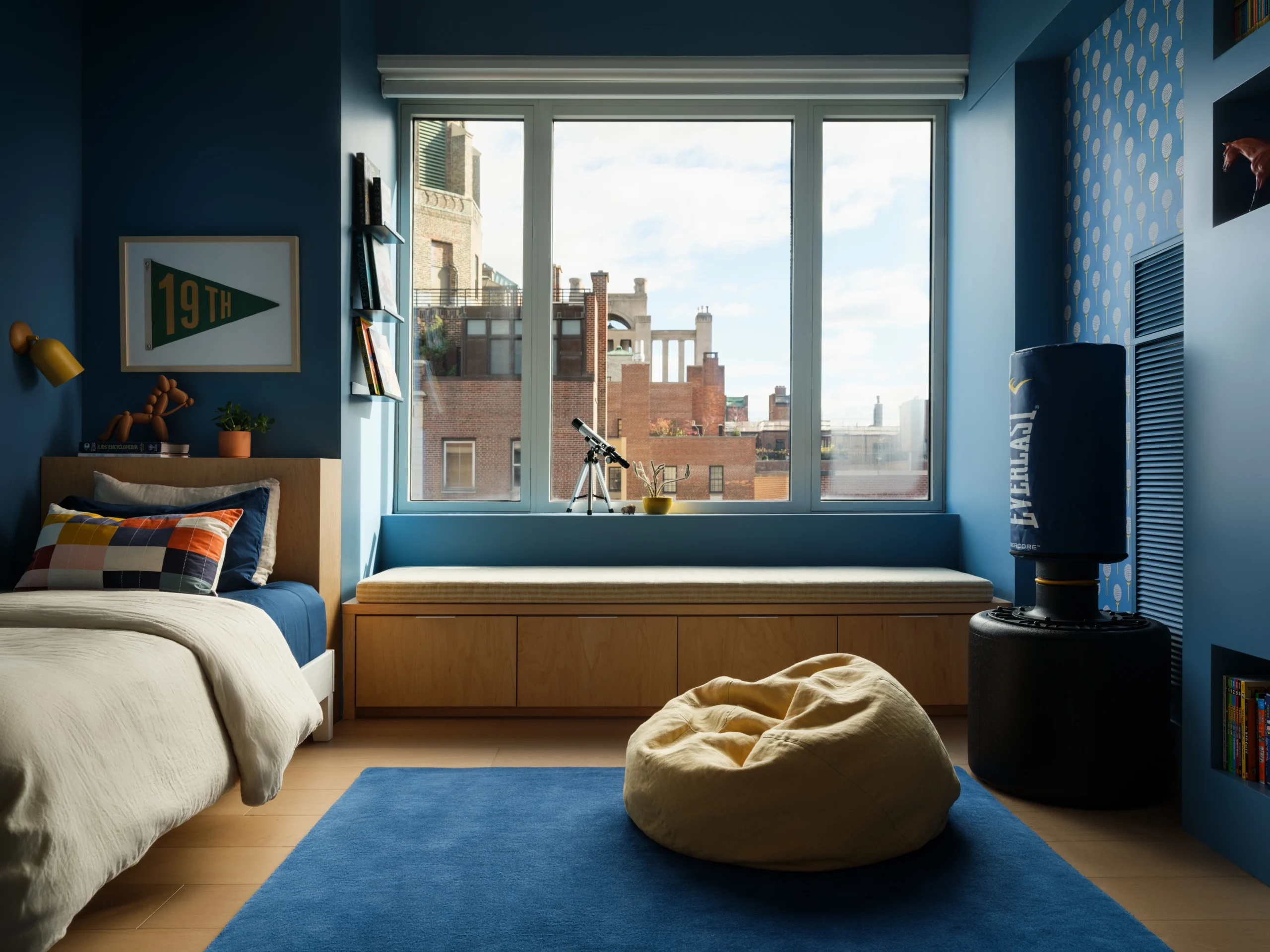 Sophisticated boys bedroom interior design in a Manhattan apartment with blue walls, built-in window seat, books, punching bag, and NYC views