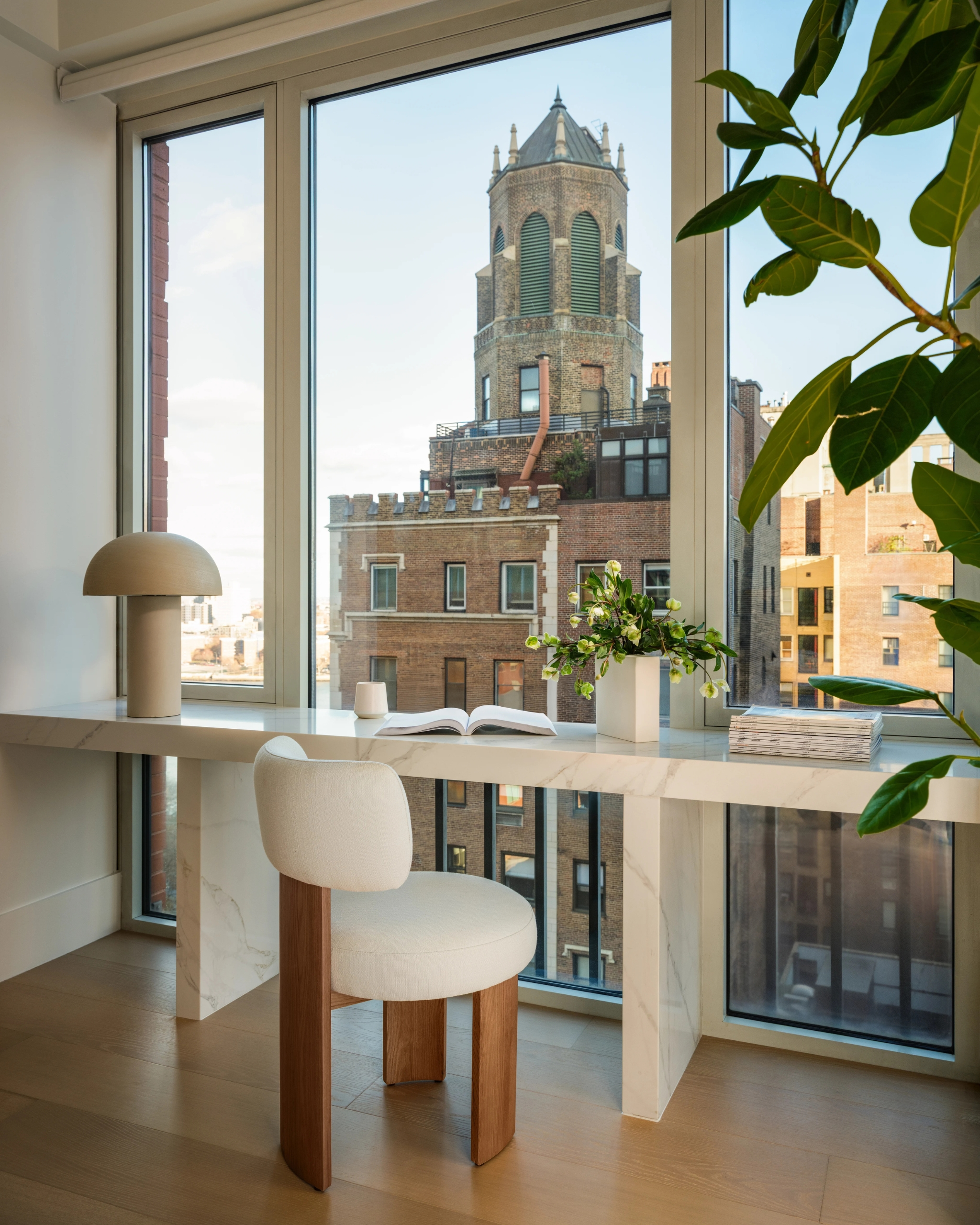 Light-filled work area in an Upper East Side NYC apartment with marble desk, soft chair, indoor plants, and Manhattan city views through large windows